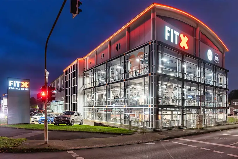 Bild: Außenansicht eines FitX Fitnessstudios. Ein großes FitX-Logo mit orangefarbenem X ist zu sehen. Das Gebäude befindet sich in einem urbanen Umfeld.