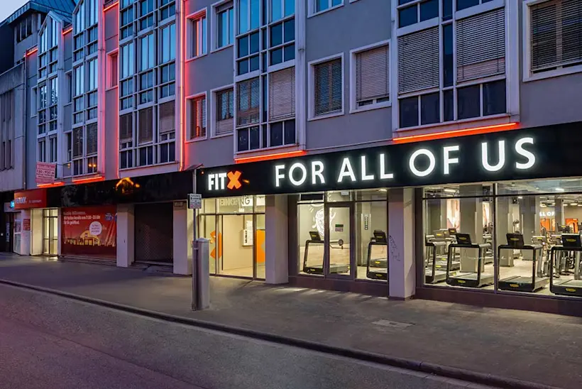 Bild: Außenansicht eines FitX Fitnessstudios. Ein großes FitX-Logo mit orangefarbenem X ist zu sehen. Das Gebäude befindet sich in einem urbanen Umfeld.