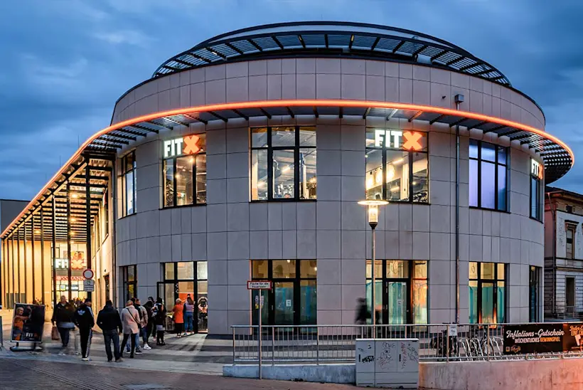 Bild: Außenansicht eines FitX Fitnessstudios. Ein großes FitX-Logo mit orangefarbenem X ist zu sehen. Das Gebäude befindet sich in einem urbanen Umfeld.