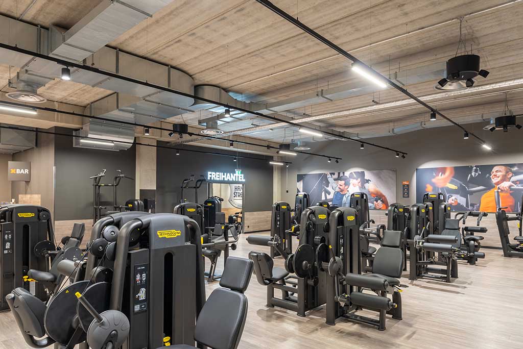 Gerätebereich im FitX Fitnessstudio Leipzig-Neulindenau mit verschiedenen Kraftgeräten. Der Raum ist im industriellen Design gestaltet, mit offenen Deckenstrukturen und hellen Böden.