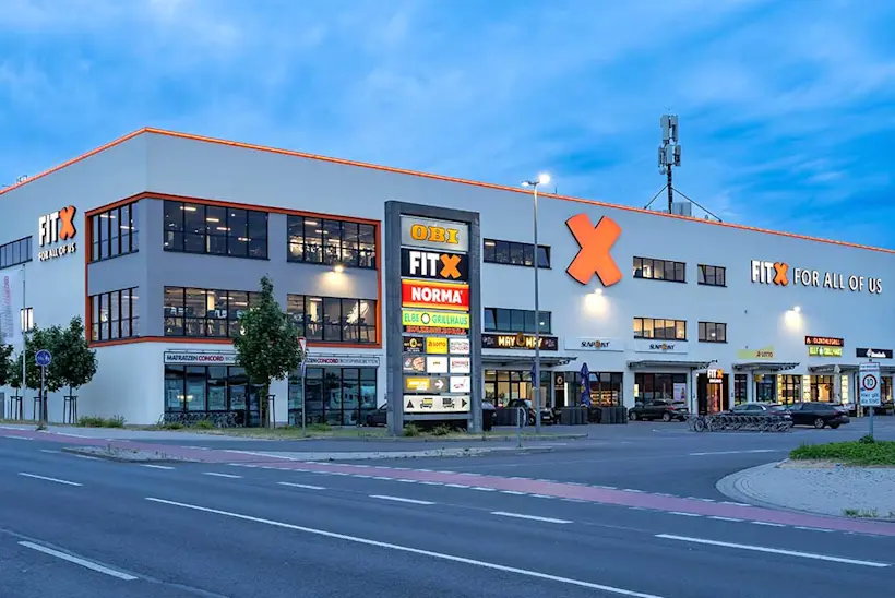 Bild: Außenansicht eines FitX Fitnessstudios. Ein großes FitX-Logo mit orangefarbenem X ist zu sehen. Das Gebäude befindet sich in einem urbanen Umfeld.