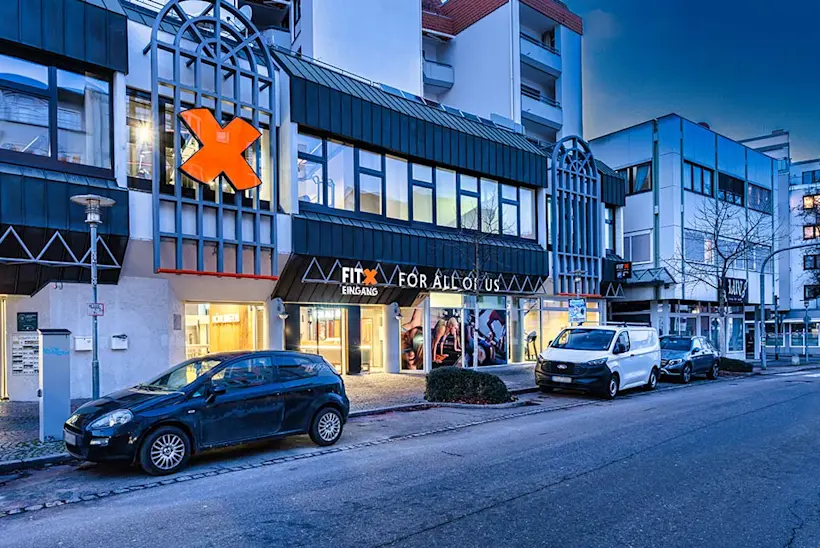 Bild: Außenansicht eines FitX Fitnessstudios. Ein großes FitX-Logo mit orangefarbenem X ist zu sehen. Das Gebäude befindet sich in einem urbanen Umfeld.