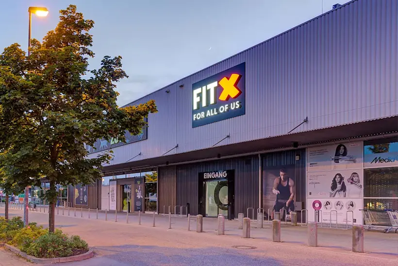 Bild: Außenansicht eines FitX Fitnessstudios. Ein großes FitX-Logo mit orangefarbenem X ist zu sehen. Das Gebäude befindet sich in einem urbanen Umfeld.