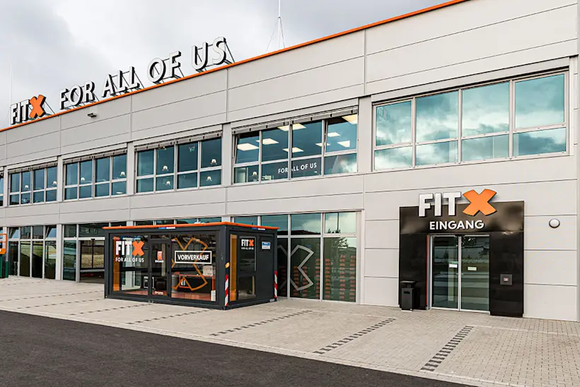 Bild: Außenansicht eines FitX Fitnessstudios. Ein großes FitX-Logo mit orangefarbenem X ist zu sehen. Das Gebäude befindet sich in einem urbanen Umfeld.