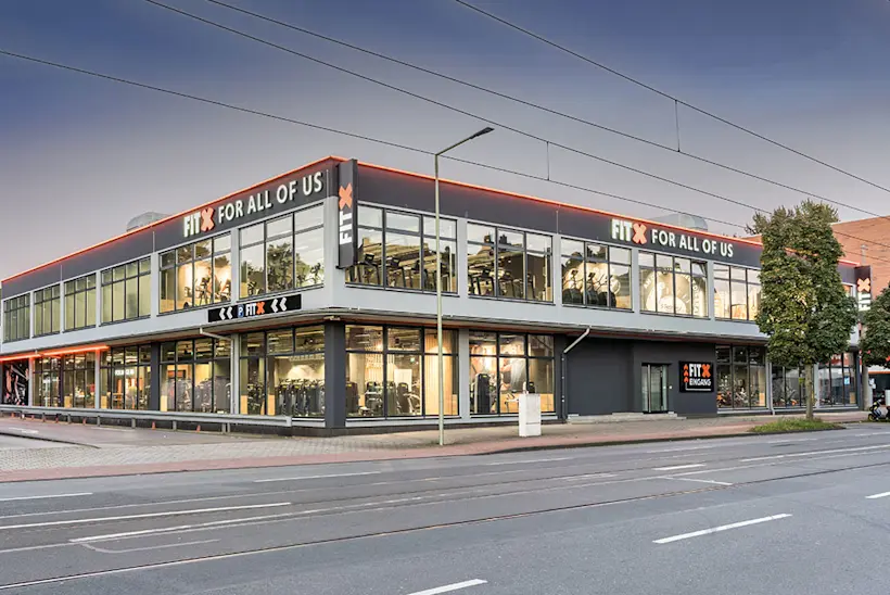 Bild: Außenansicht eines FitX Fitnessstudios. Ein großes FitX-Logo mit orangefarbenem X ist zu sehen. Das Gebäude befindet sich in einem urbanen Umfeld.