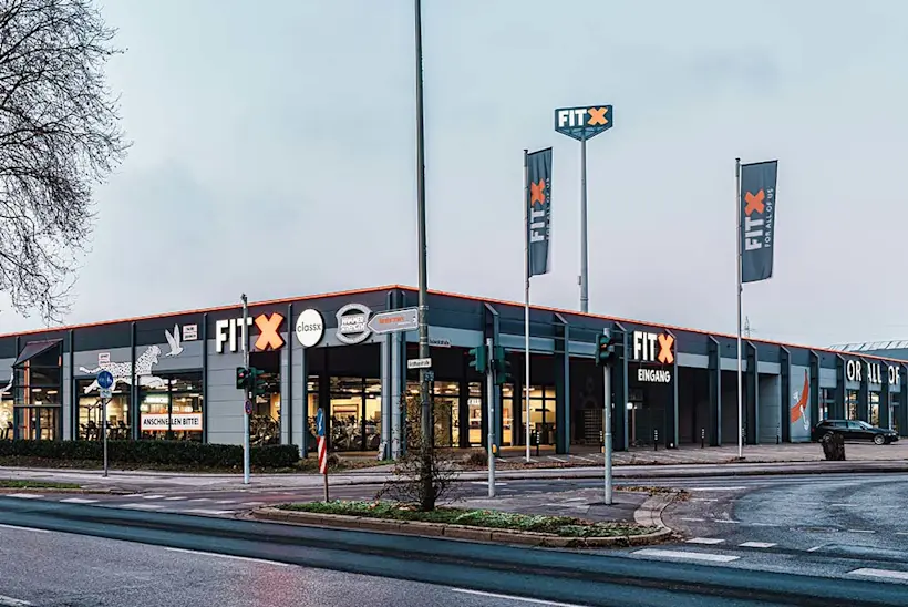 Bild: Außenansicht eines FitX Fitnessstudios. Ein großes FitX-Logo mit orangefarbenem X ist zu sehen. Das Gebäude befindet sich in einem urbanen Umfeld.