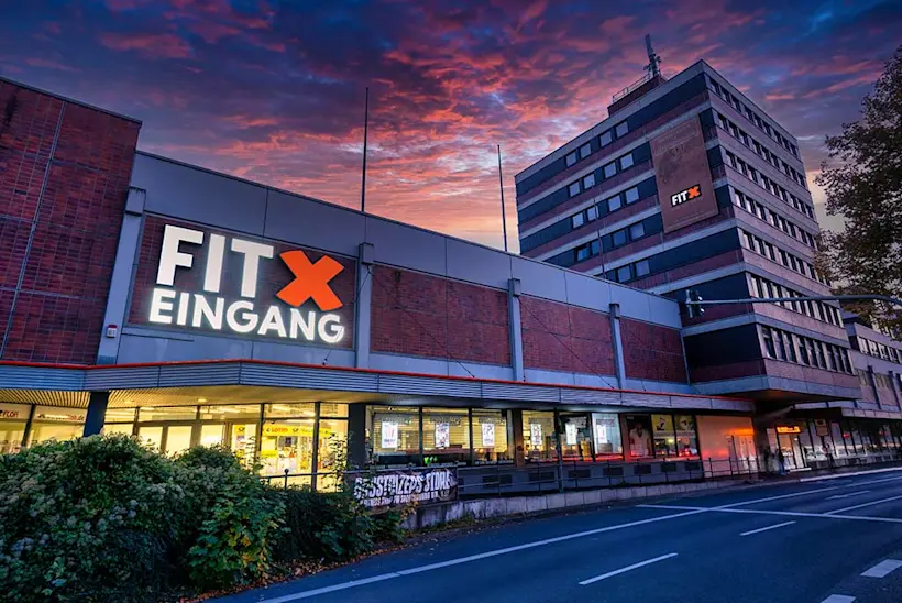 Bild: Außenansicht eines FitX Fitnessstudios. Ein großes FitX-Logo mit orangefarbenem X ist zu sehen. Das Gebäude befindet sich in einem urbanen Umfeld.