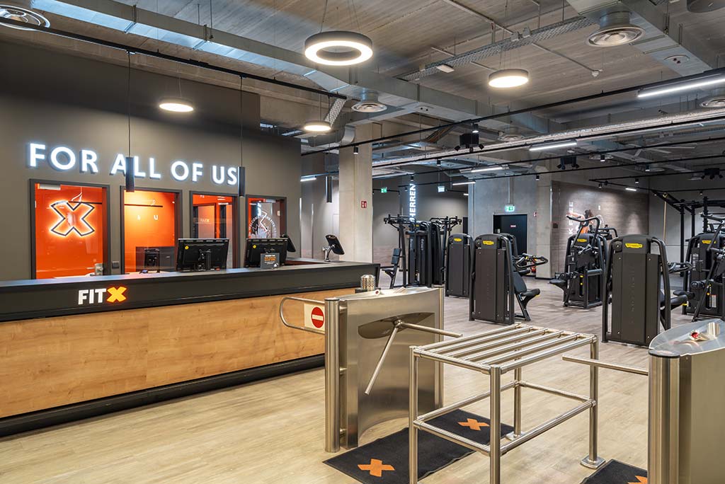 Empfangsbereich des FitX Fitnessstudios Remscheid-Süd mit einer holzverkleideten Theke, einem Drehkreuz am Eingang und dem leuchtenden Schriftzug „FOR ALL OF US“ an der Wand.