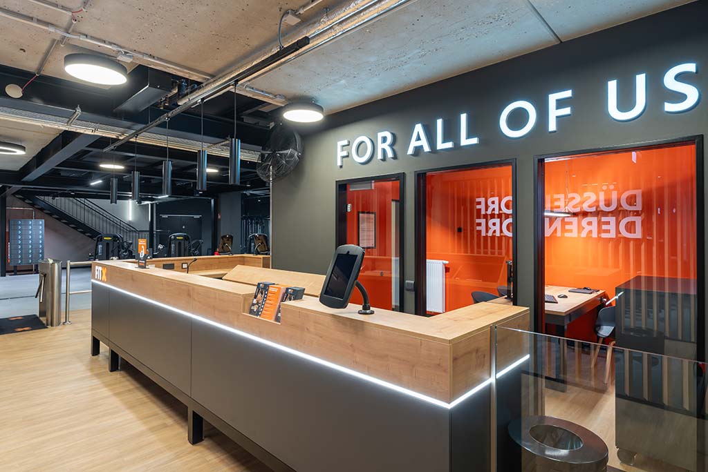 Empfangsbereich des FitX Fitnessstudios Düsseldorf-Derendorf mit einer holzverkleideten Theke, einem Drehkreuz am Eingang und dem leuchtenden Schriftzug „FOR ALL OF US“ an der Wand.
