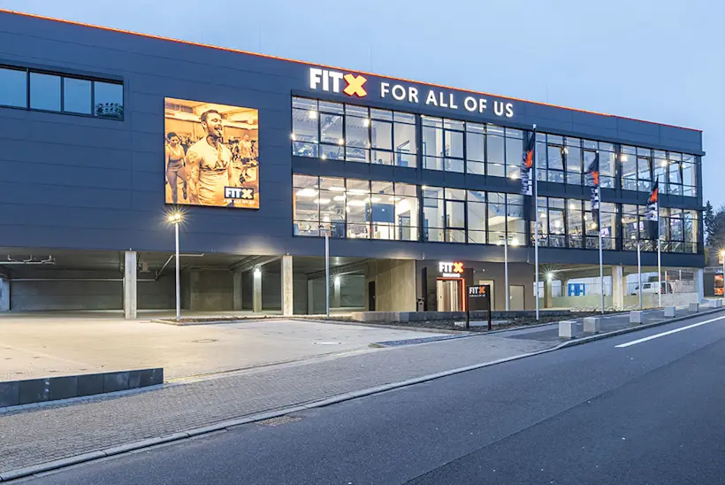 Bild: Außenansicht eines FitX Fitnessstudios. Ein großes FitX-Logo mit orangefarbenem X ist zu sehen. Das Gebäude befindet sich in einem urbanen Umfeld.