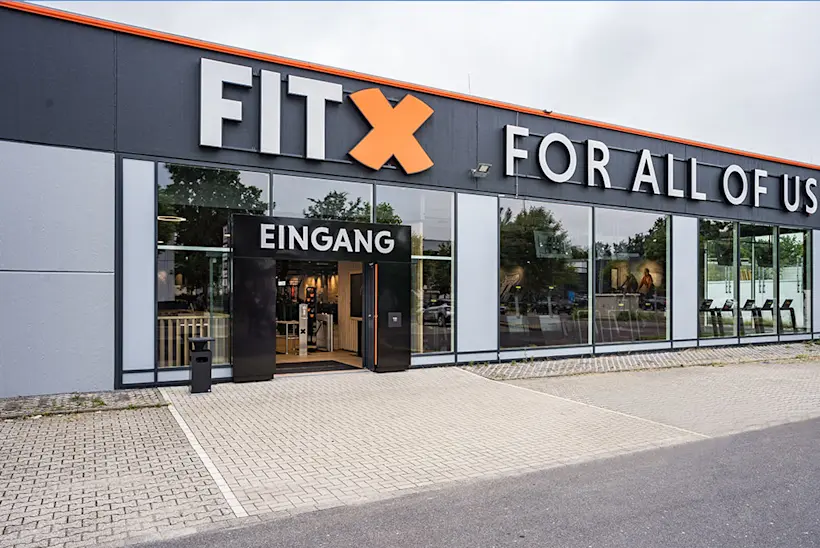Bild: Außenansicht eines FitX Fitnessstudios. Ein großes FitX-Logo mit orangefarbenem X ist zu sehen. Das Gebäude befindet sich in einem urbanen Umfeld.