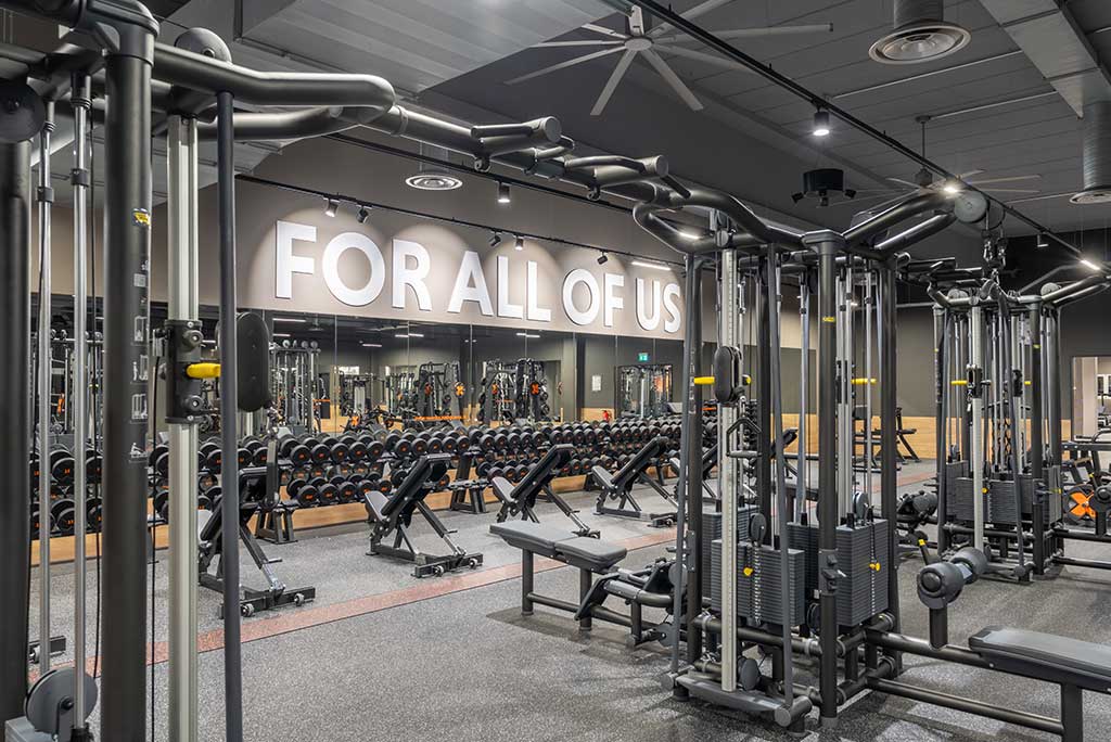 Weitläufiger Freihantelbereich mit verschiedenen Kraftgeräten im FitX Fitnessstudio Nürnberg-Zabo. An der Seite sind Kurzhanteln vor einer Spiegelwand aufgereiht. An der oberen Wand prangt der Schriftzug „FOR ALL OF US“.