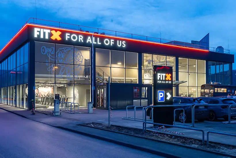Bild: Außenansicht eines FitX Fitnessstudios. Ein großes FitX-Logo mit orangefarbenem X ist zu sehen. Das Gebäude befindet sich in einem urbanen Umfeld.