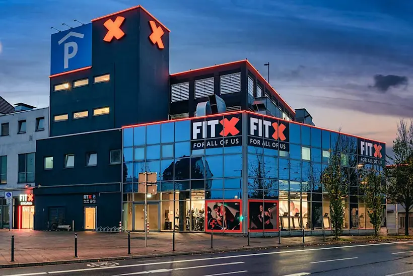 Bild: Außenansicht eines FitX Fitnessstudios. Ein großes FitX-Logo mit orangefarbenem X ist zu sehen. Das Gebäude befindet sich in einem urbanen Umfeld.