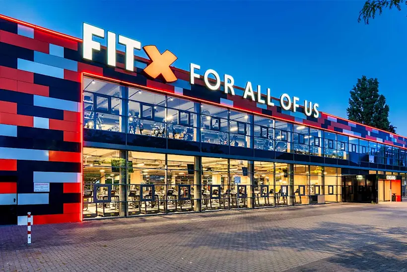Bild: Außenansicht eines FitX Fitnessstudios. Ein großes FitX-Logo mit orangefarbenem X ist zu sehen. Das Gebäude befindet sich in einem urbanen Umfeld.