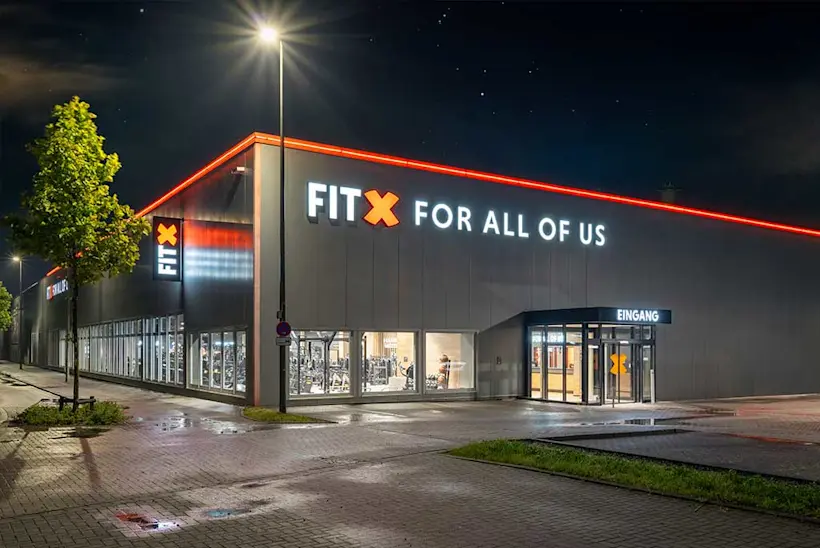 Bild: Außenansicht eines FitX Fitnessstudios. Ein großes FitX-Logo mit orangefarbenem X ist zu sehen. Das Gebäude befindet sich in einem urbanen Umfeld.