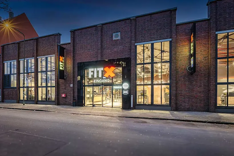 Bild: Außenansicht eines FitX Fitnessstudios. Ein großes FitX-Logo mit orangefarbenem X ist zu sehen. Das Gebäude befindet sich in einem urbanen Umfeld.