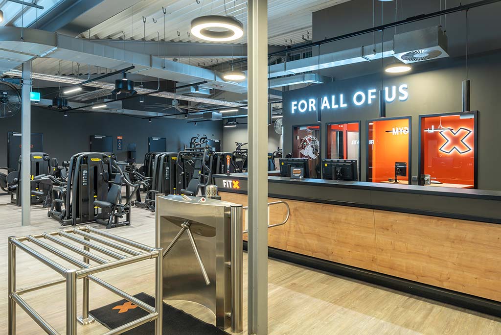 Empfangsbereich des FitX Fitnessstudios Neuss-Innenstadt mit einer holzverkleideten Theke, einem Drehkreuz am Eingang und dem leuchtenden Schriftzug „FOR ALL OF US“ an der Wand.