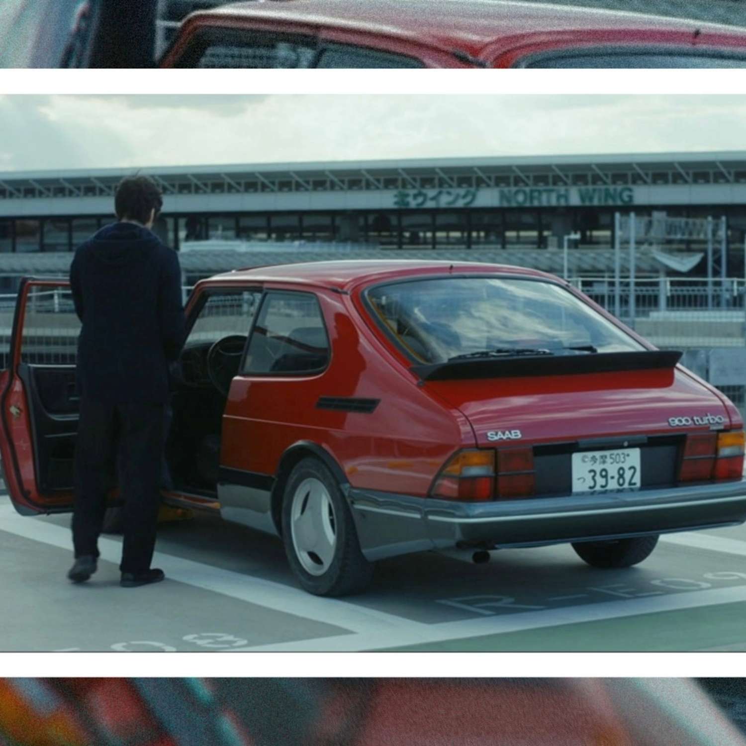 Still from Drive My Car, with Yūsuke standing in the open door of a red Saab 900 Turbo