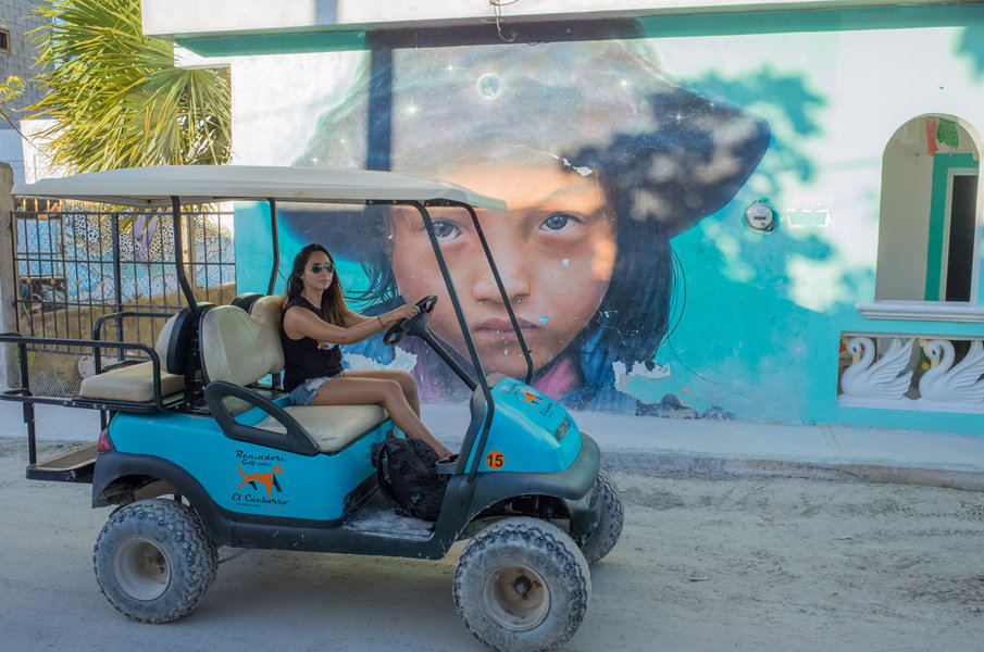 A young woman driving a rental golf cart next to the Isla Holbox wooden pier on a sunny day.