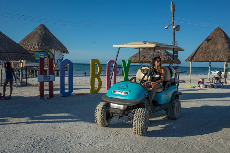 A young woman driving a rental golf cart next to the Isla Holbox wooden pier on a sunny day.