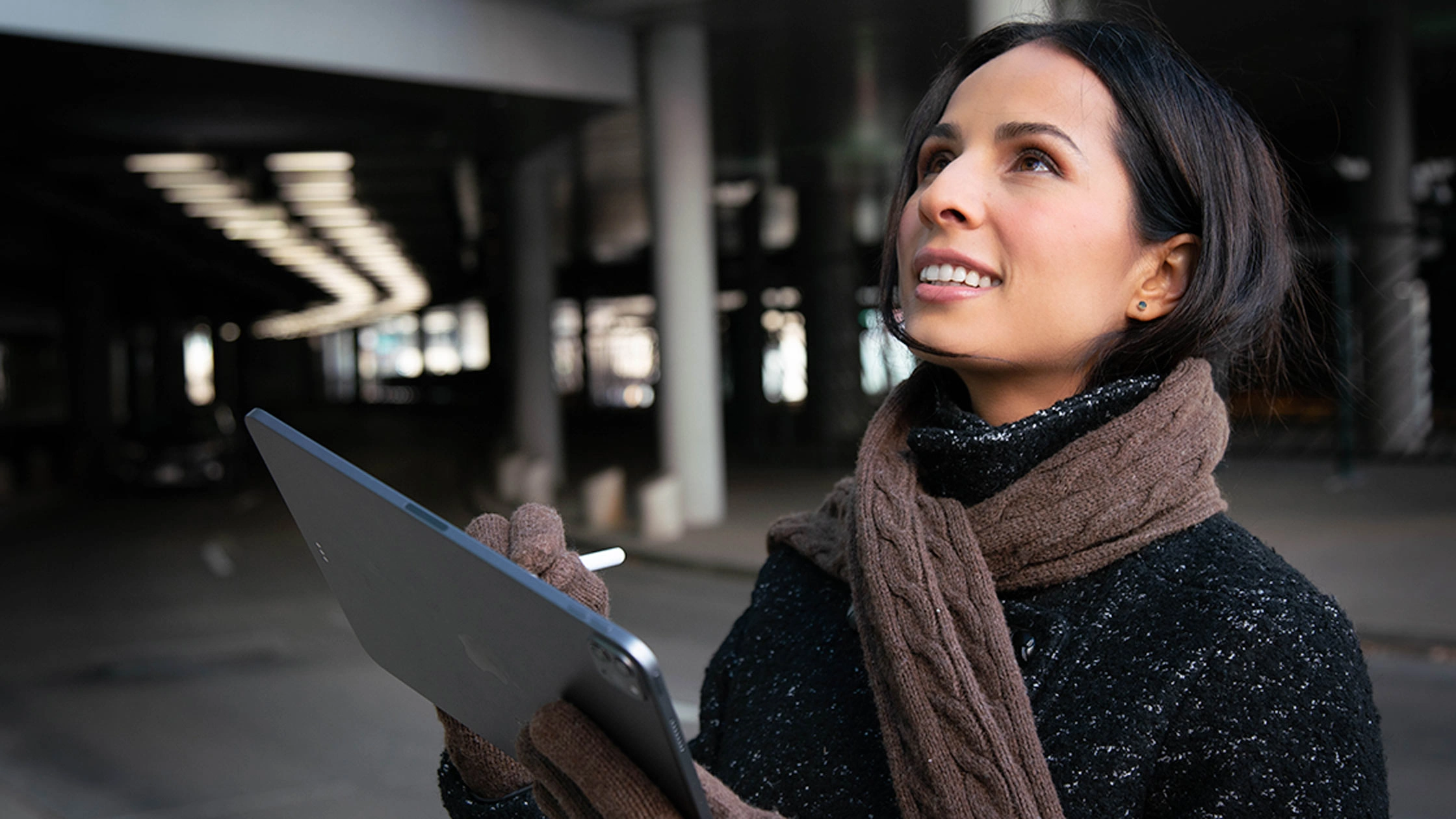 A woman in winter attire looks up while using a stylus on a Trimble rugged tablet in a city setting.