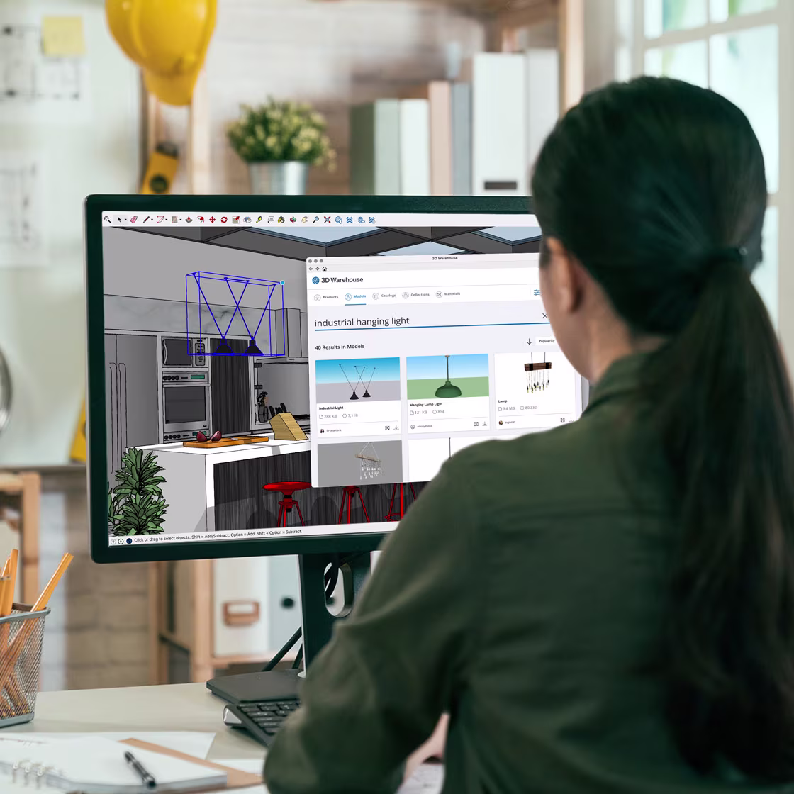 Eine Person sitzt an einem Schreibtisch und arbeitet an einem Computer, auf dessen Bildschirm 3D Warehouse und SketchUp geöffnet sind.