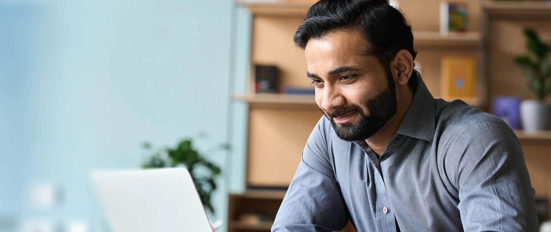 A professional man with a beard looking focused and smiling slightly while working on a laptop in a bright, modern office setting