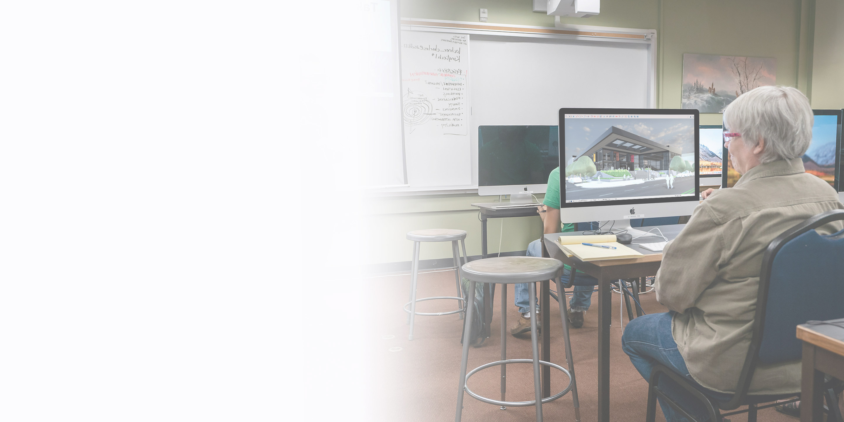 A student works in SketchUp in a classroom on a desktop computer while a teacher stands at the whiteboard.