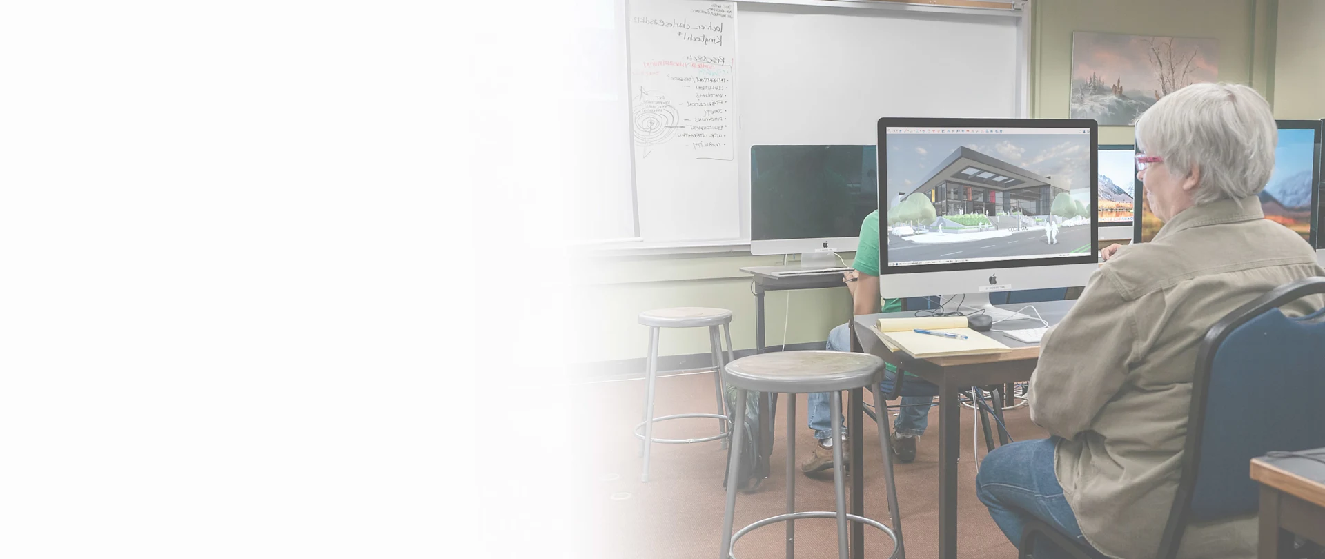 A student works in SketchUp in a classroom on a desktop computer while a teacher stands at the whiteboard.
