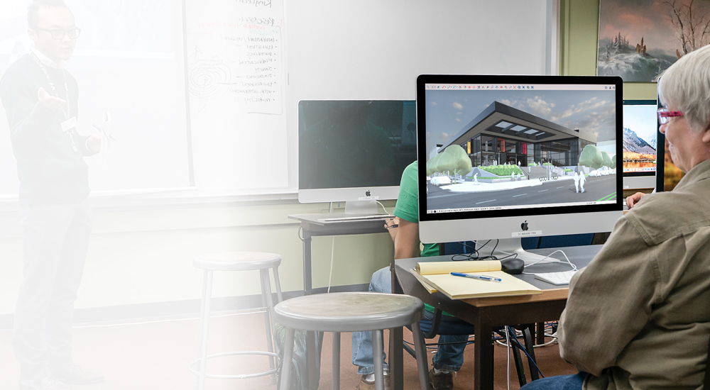 A student works in SketchUp in a classroom on a desktop computer while a teacher stands at the whiteboard.