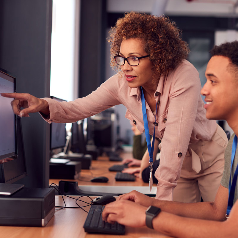 一名學生坐在電腦桌前，老師則站在一旁指著螢幕