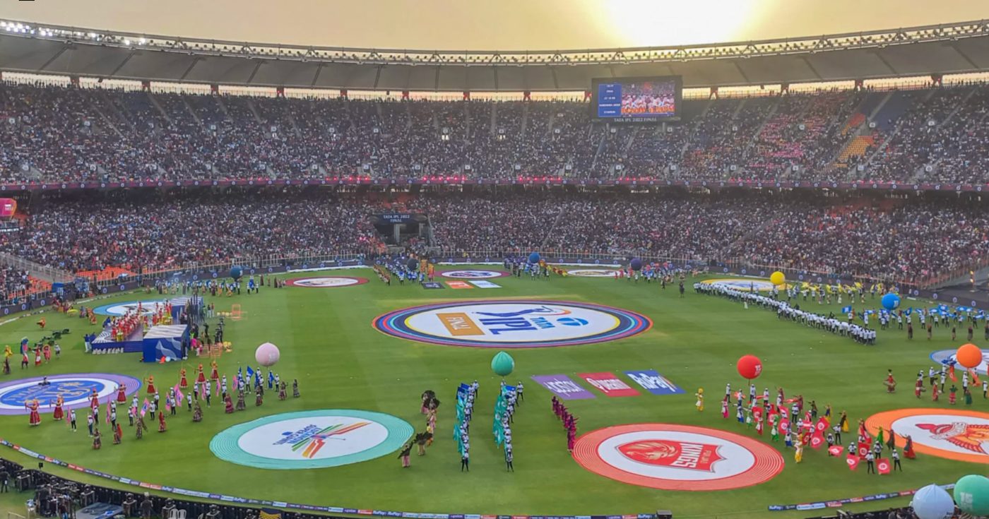 A packed IPL stadium during a night match, floodlights washing the field, large crowd of spectators in team colours