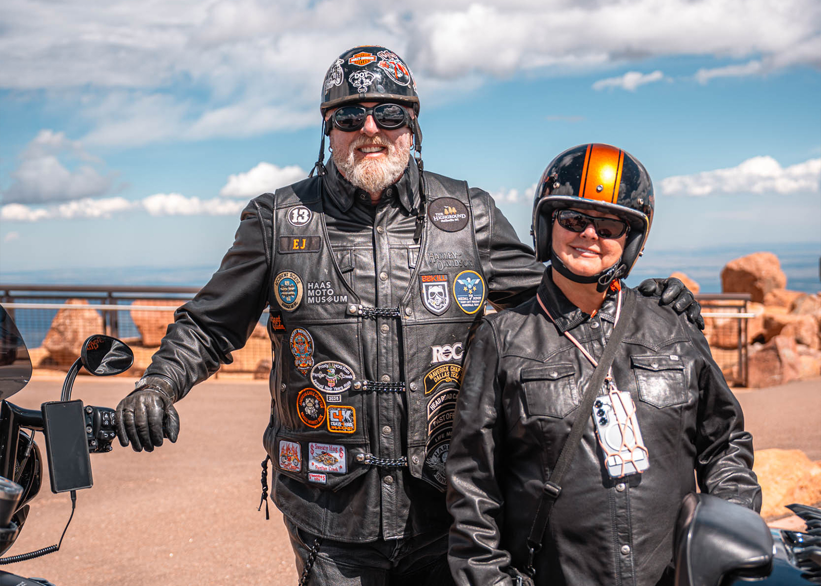 Two H.O.G. members in leather jackets with patches and helmets smile for a photo at the summit of Pikes Peak.