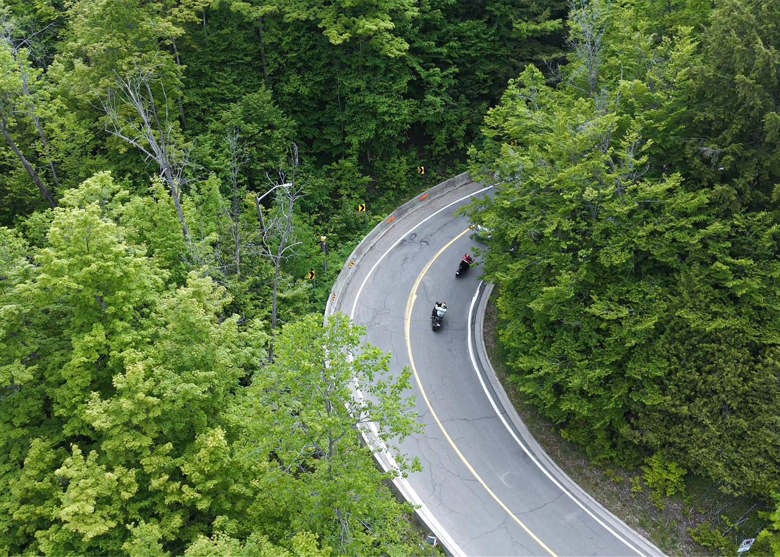 Aerial view of motorcyclists leaning into a sharp forested curve on a winding road.