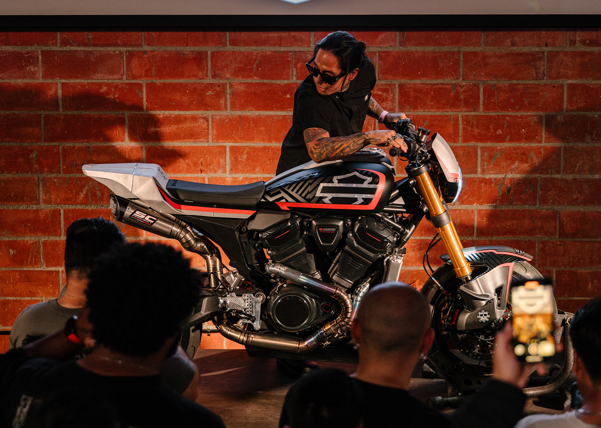 Sean Guardado of SMCO presents a custom Harley-Davidson motorcycle on stage, admired by a crowd capturing photos.