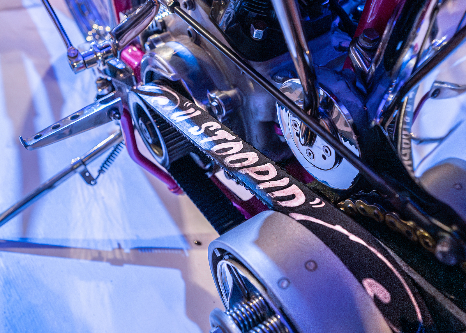 A belt drive on a motorcycle reads "U STOOPID" with hand-painted lettering 