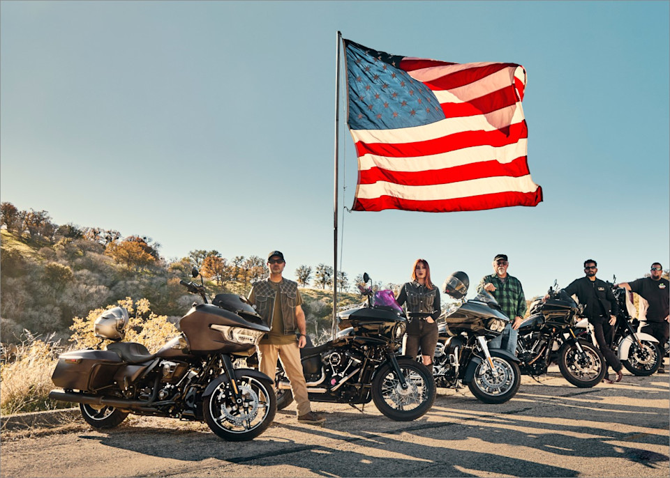 Grupo de motociclistas con motocicletas bajo una gran bandera estadounidense al aire libre