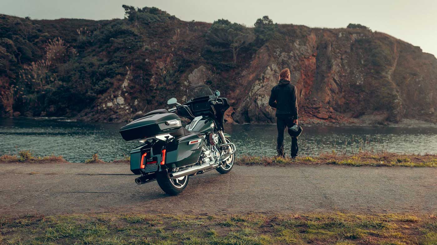 Motorcycle parked on road with riding standing looking into the distance with water and mountains in the background