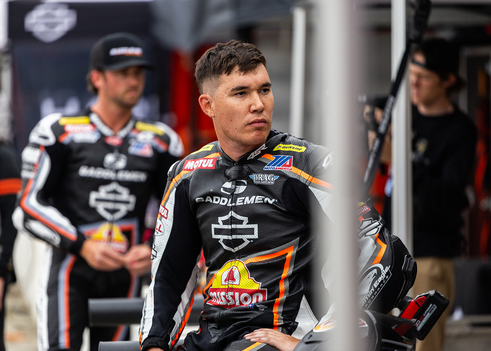 One of the racers looks sternly into the distance, with his teammates standing behind him slightly out of focus. 