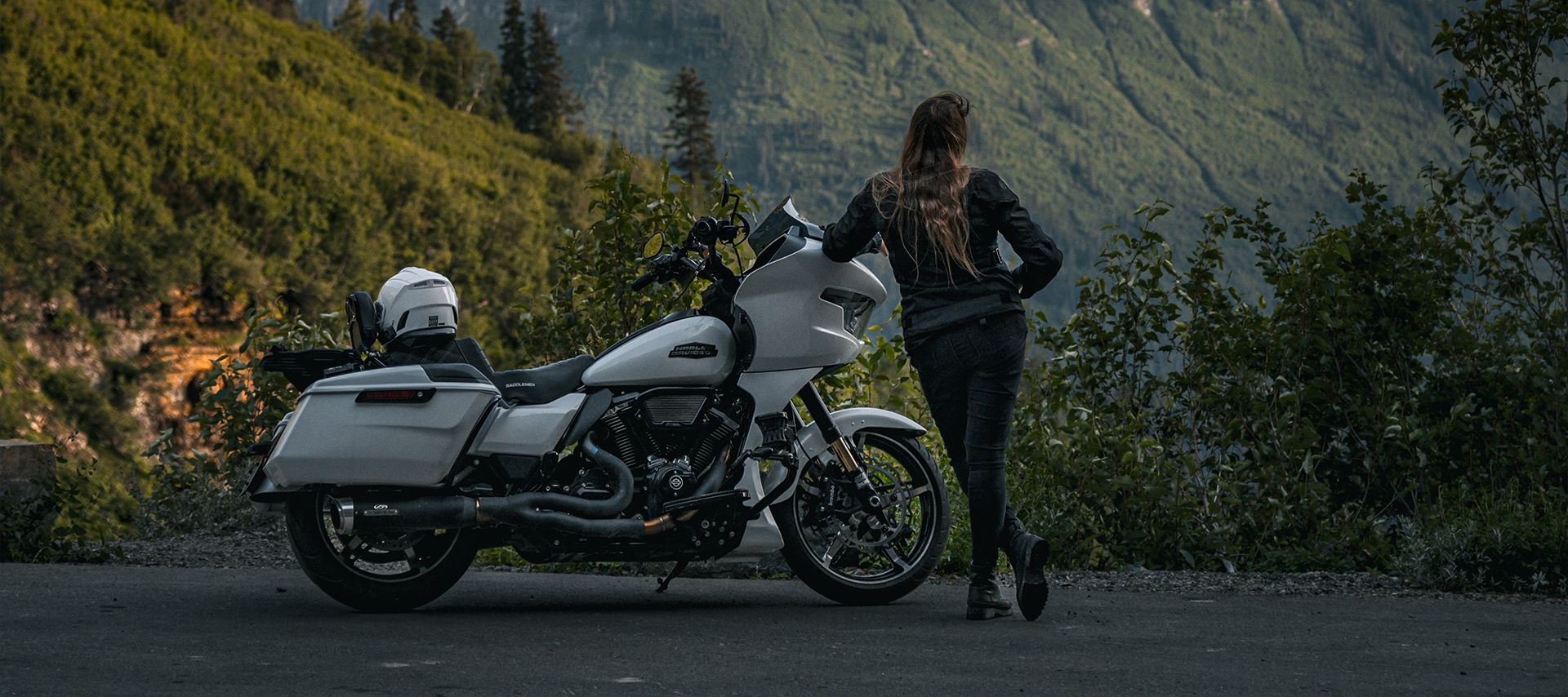 The author stands next to a white Harley-Davidson Road Glide motorcycle on a mountain roadside with her back to the camera.
