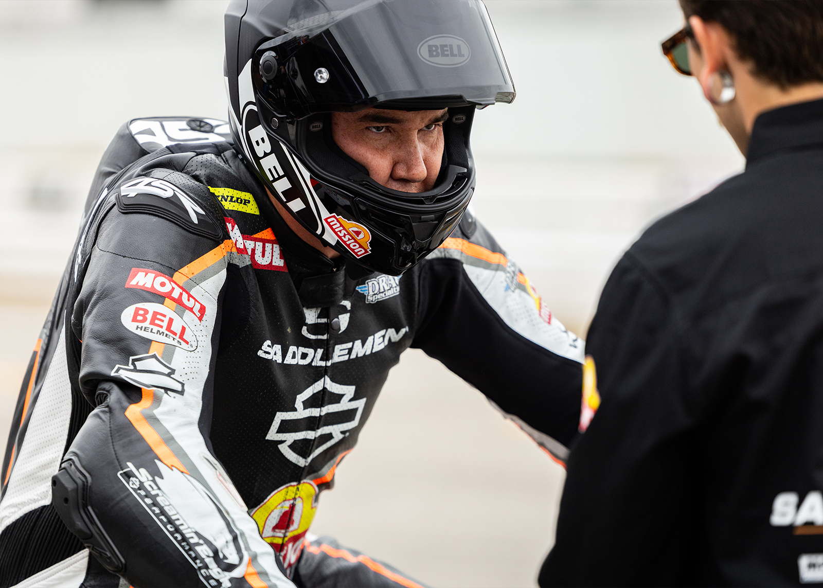 A closeup of a rider before the race with his helmet is up talking to another member of the race team. He looks focused and prepared.