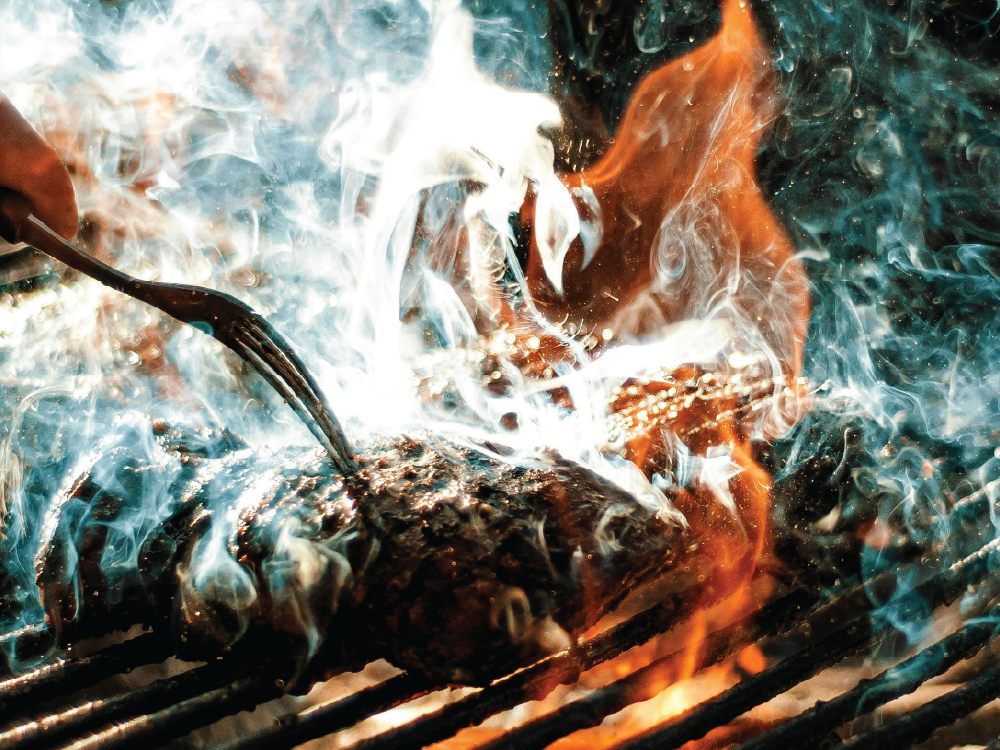 Meat grilling over open flame with smoke and tongs