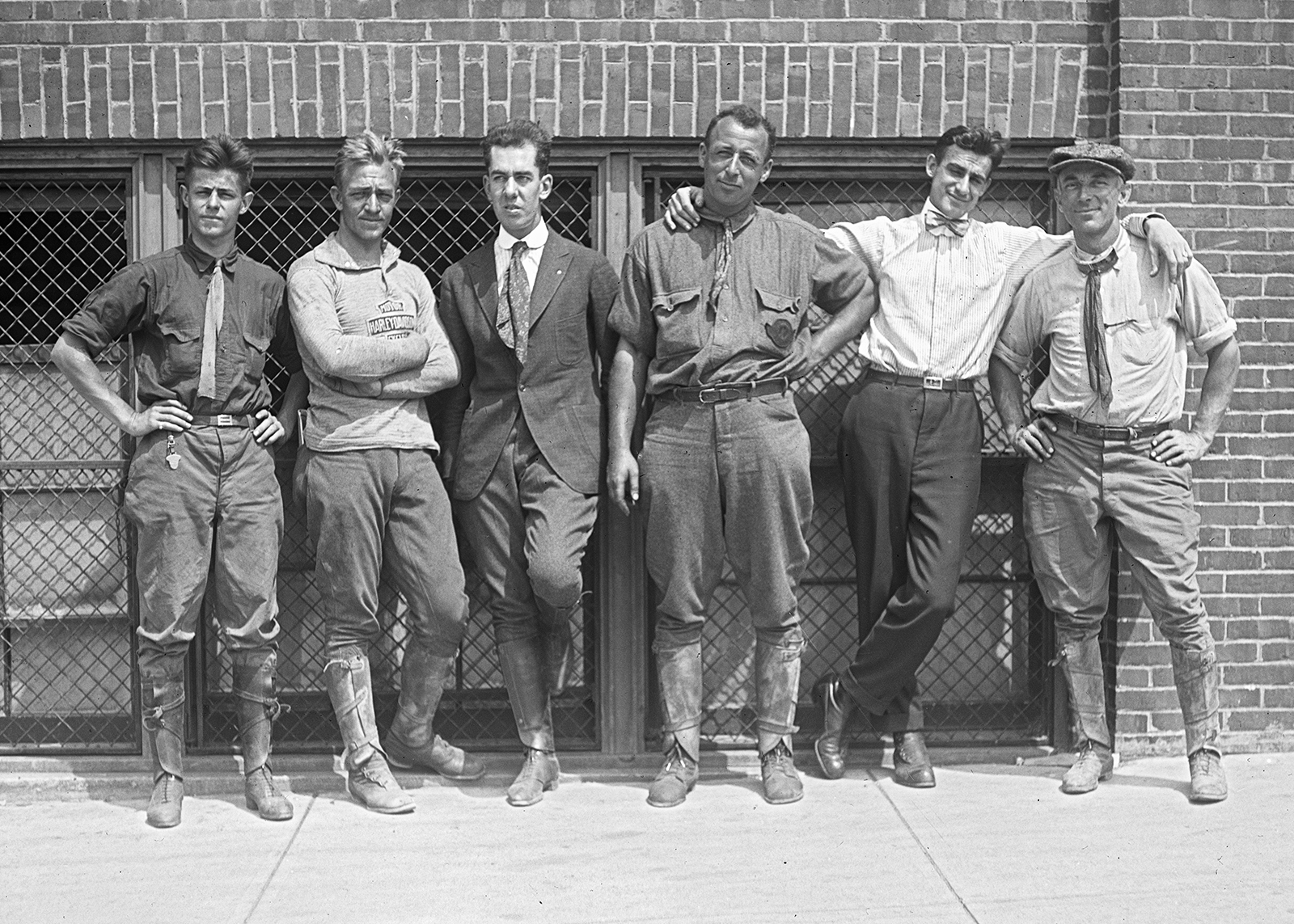 Vintage black-and-white photo of six men in Harley-Davidson gear, posing by a brick building.