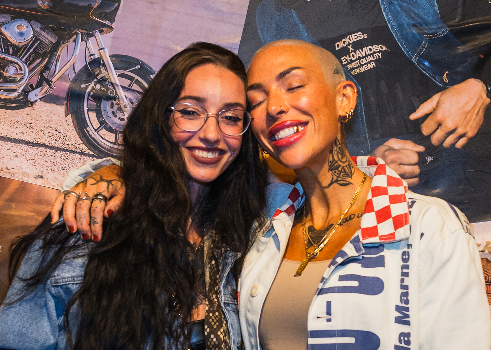 Two women, one with black hair and the other with a shaved head, smile with their heads leaned together in front of a wall of images of the H-D x Dickies collab.