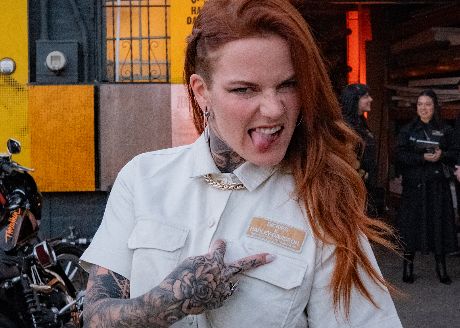A woman with red hair sticks her tongue out and points to her HD x Dickies patch on her white shirt.