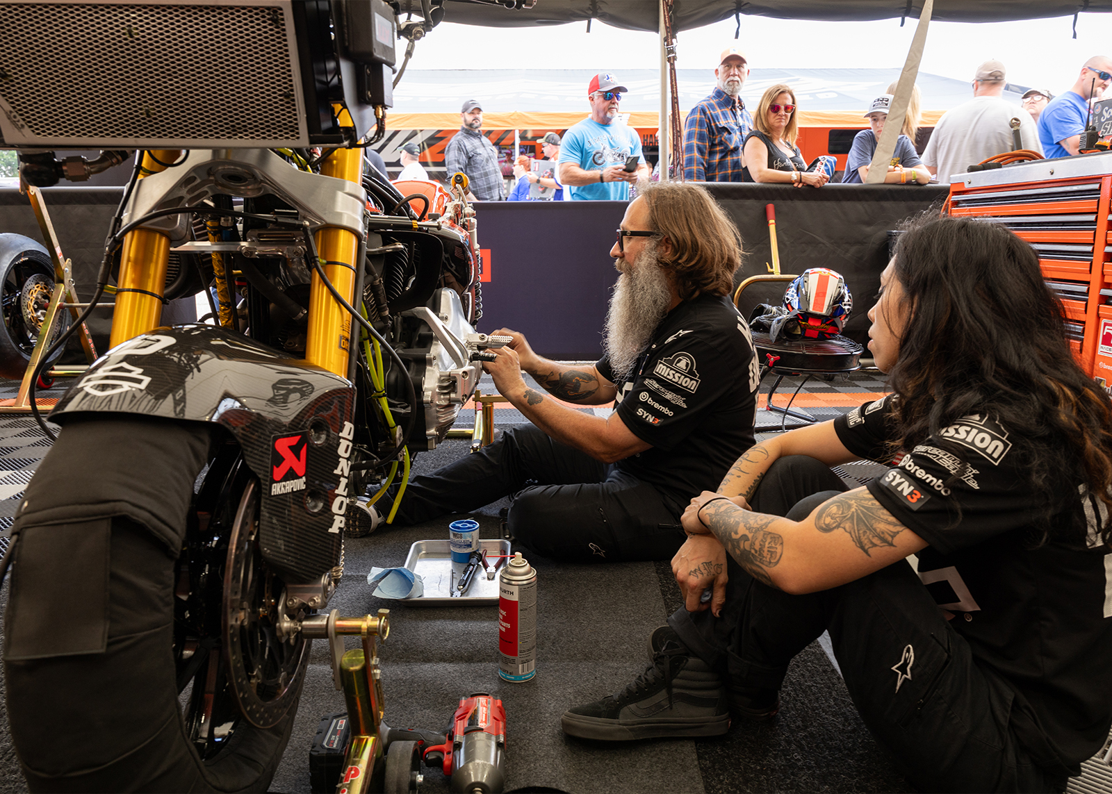 Two HD racing team members sit down and work on one of the bikes, with a crowd of onlookers behind them. 