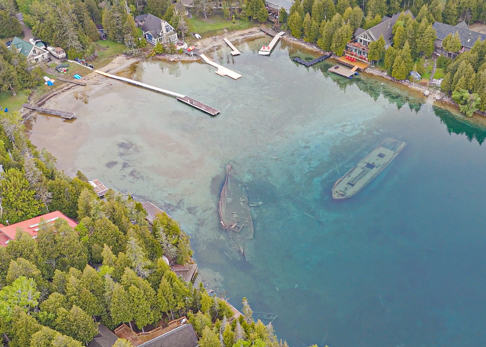 Aerial view of shipwrecks visible under clear water near docks and lakeside homes surrounded by trees.
