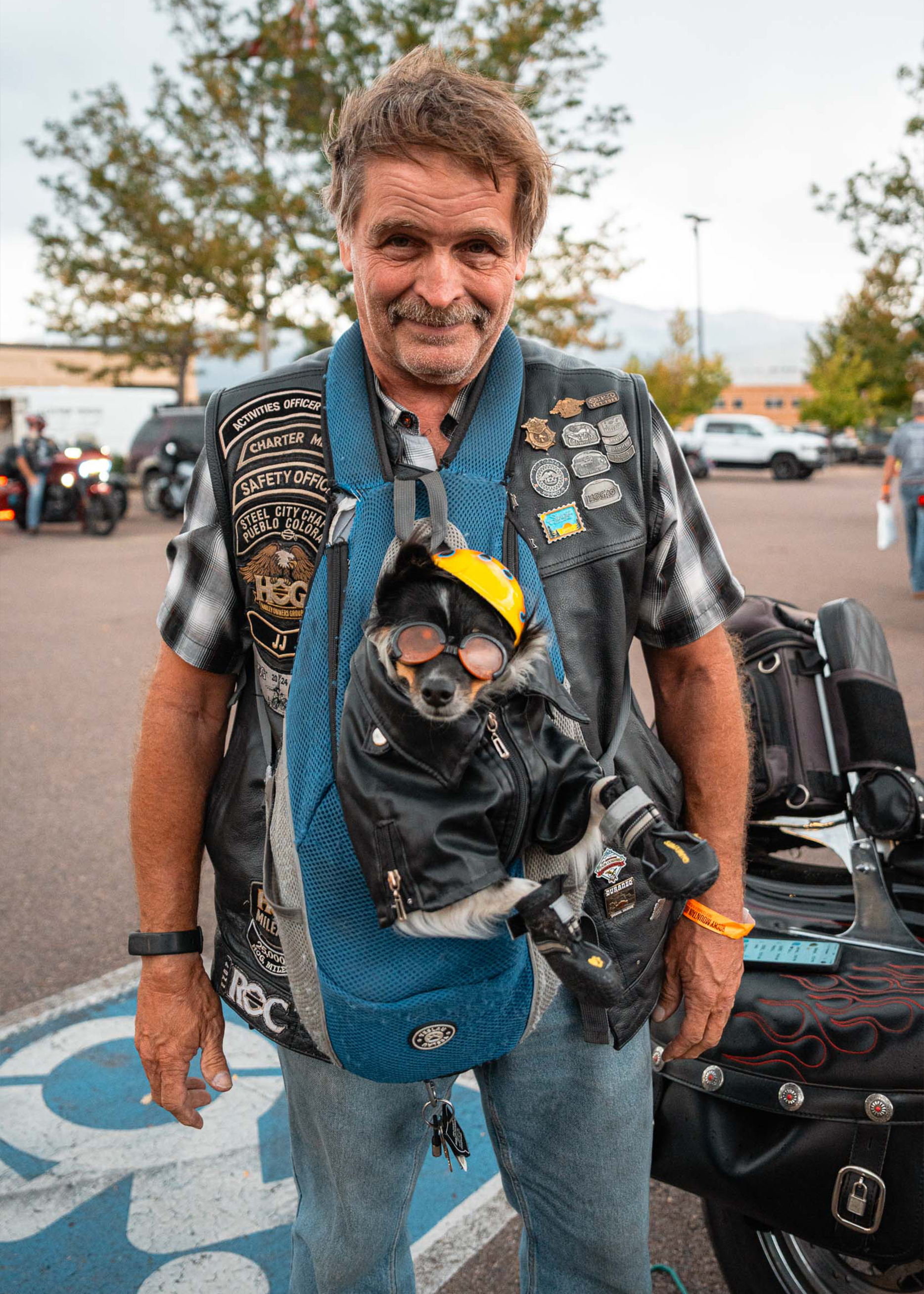 A H.O.G. member poses next to his motorcycle, carrying a small dog wearing a helmet, leather jacket, boots, and sunglasses in a pack on his front.