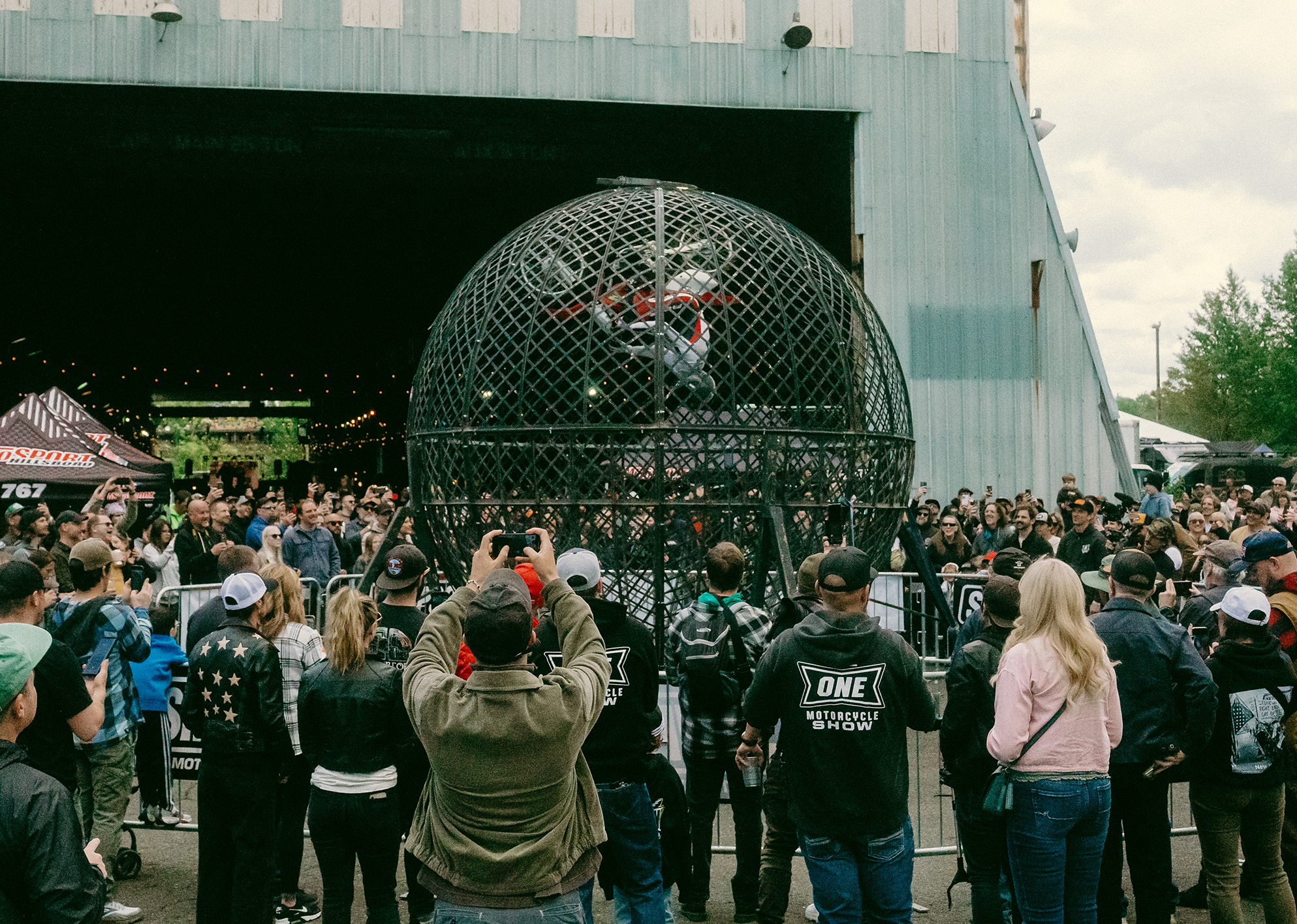 A rider and his bike are moving upside-down in a metal spherical cage. A large crowd of people watch in amazement, many holding up their phones to capture the spectacle.